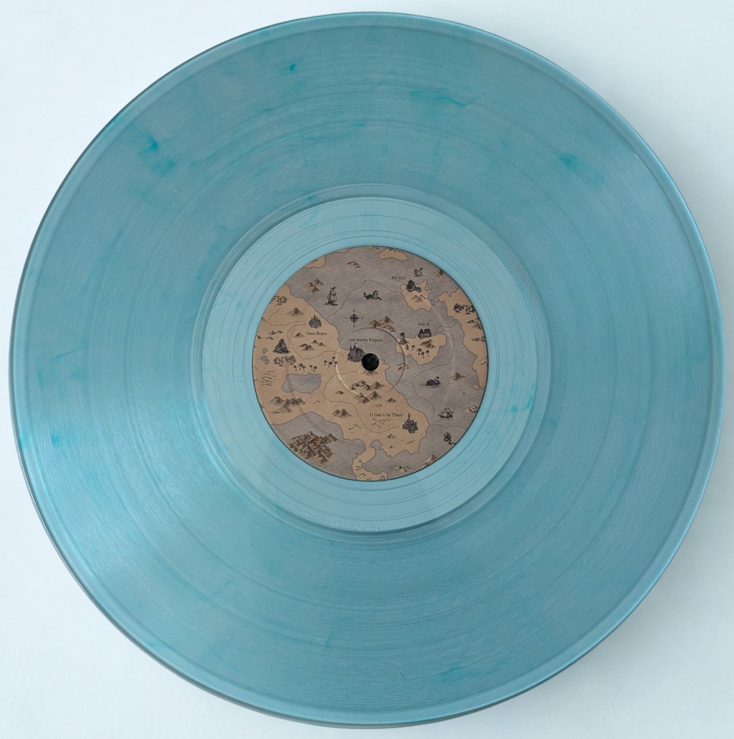 A translucent light blue vinyl record with a detailed fantasy map-style label in the center, photographed from directly above on a clean white background.