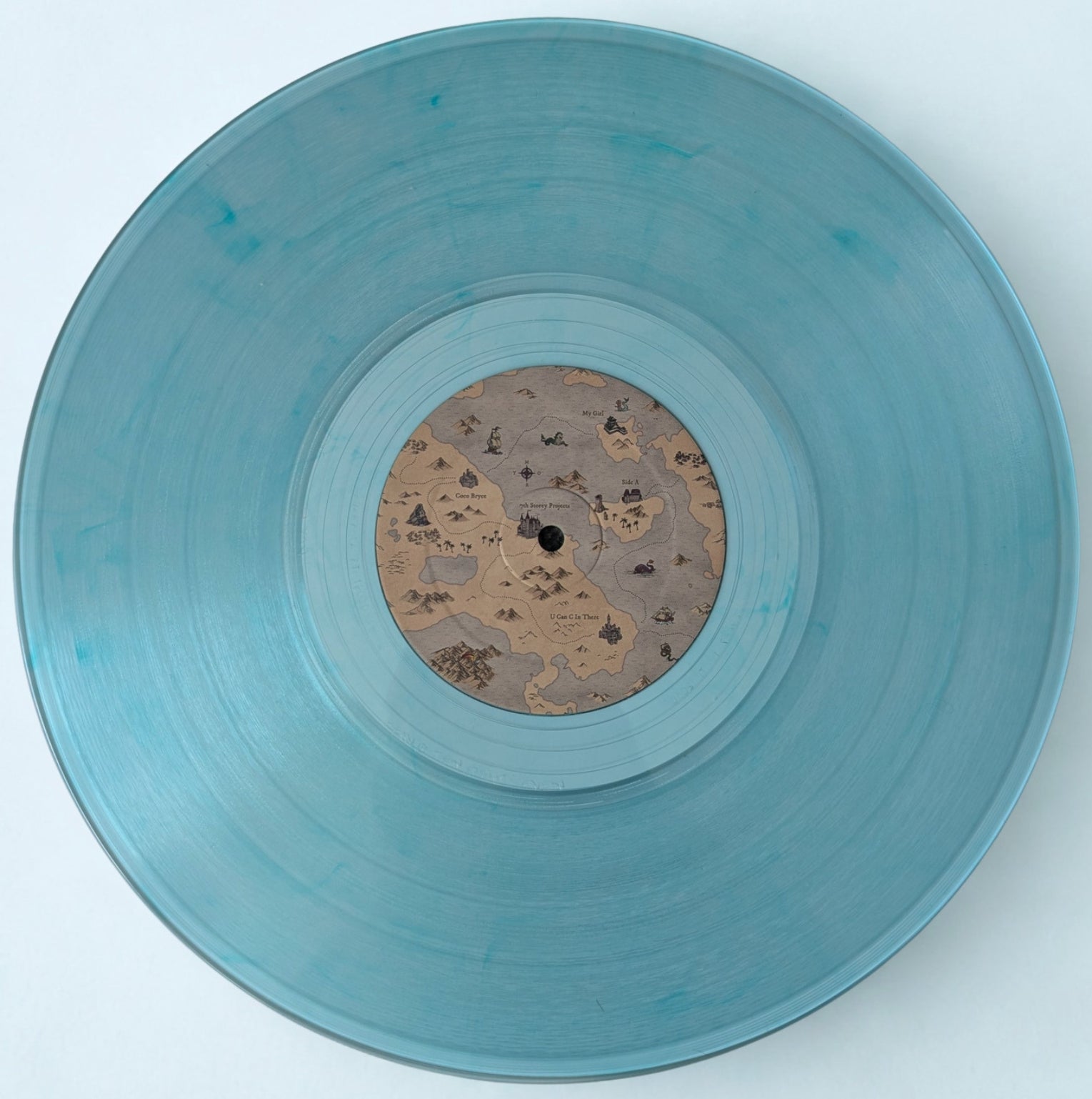 A translucent light blue vinyl record with a detailed fantasy map-style label in the center, photographed from directly above on a clean white background.